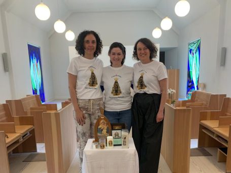 Três mulheres de camisetas brancas posam em uma mesa em um espaço iluminado.